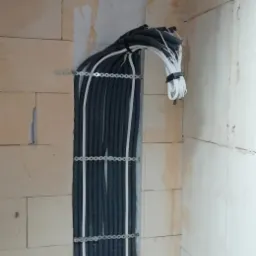 Pionowy układ peszli z przewodami elektrycznymi, przymocowany metalowymi uchwytami do ściany z bloczków, widoczne przewody wychodzące górą.