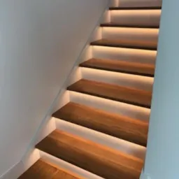 Drewniane schody z podświetleniem LED wzdłuż stopni, ciepłe światło, białe ściany i listwy przypodłogowe.