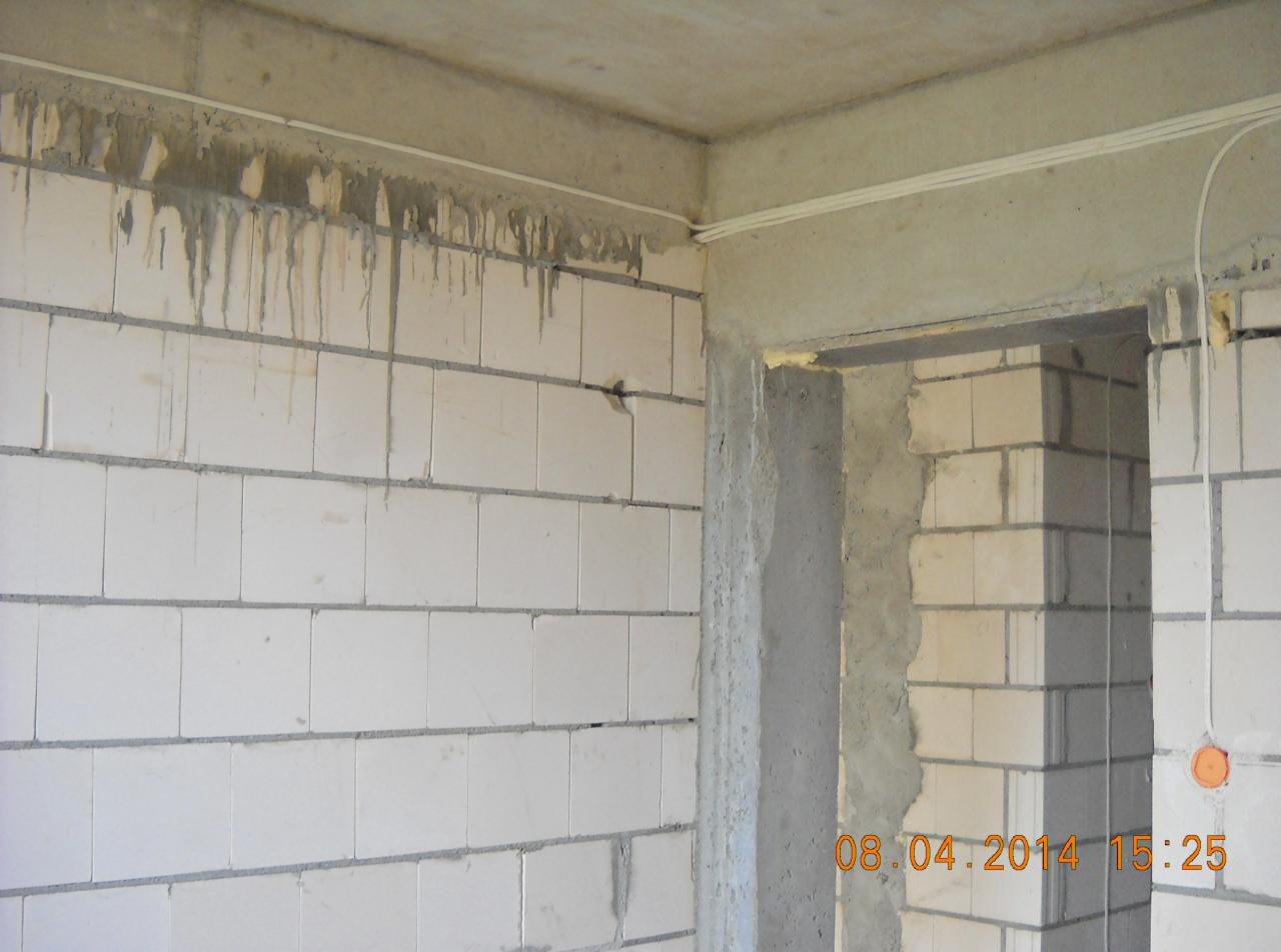 Surowy stan wnętrza: ściany z białych bloczków, betonowy strop z widocznymi kablami elektrycznymi, niedokończone obramowanie drzwi, ślady zaprawy na ścianie.