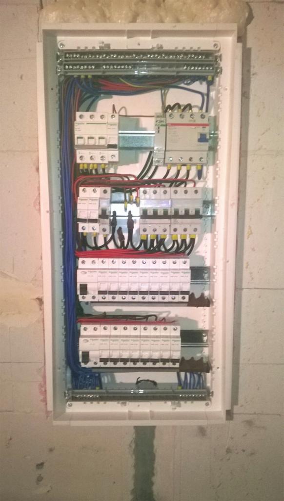 Rozdzielnica elektryczna z bezpiecznikami, przewodami i listwami zaciskowymi zamontowana w białej obudowie na surowej ścianie z betonu komórkowego.