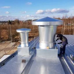 Pracownik w czarnej kurtce montuje metalową nasadę wentylacyjną z daszkiem na dachu z blachy trapezowej; w tle widoczne inne nasady, drzewa i zabudowania.