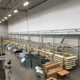 Wysoki kąt widzenia na magazyn z elementami instalacji wentylacyjnej, w tym rury, kanały i komponenty składowane na półkach, z widocznymi elementami systemu wentylacyjnego pod sufitem.