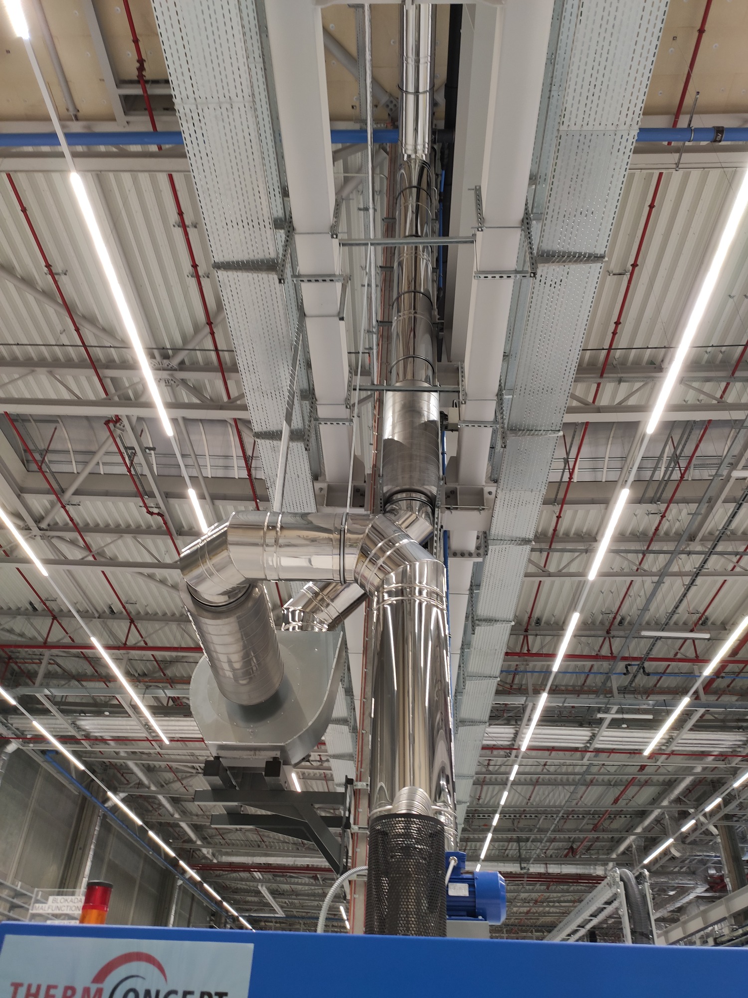Widok z dołu na system rur wentylacyjnych ze stali nierdzewnej, zamontowany pod sufitem hali produkcyjnej, z widocznymi elementami mocującymi i oświetleniem LED.