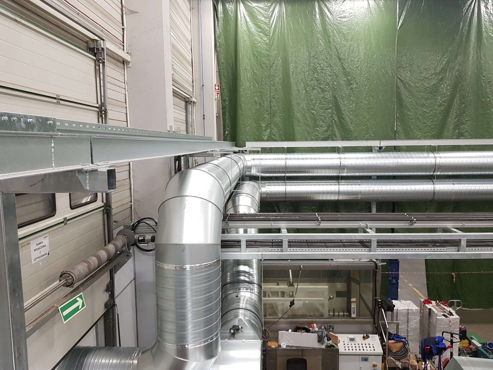 Metalowe rury wentylacyjne, zawieszone pod sufitem hali przemysłowej, z widocznym systemem wsporników i zieloną plandeką w tle.