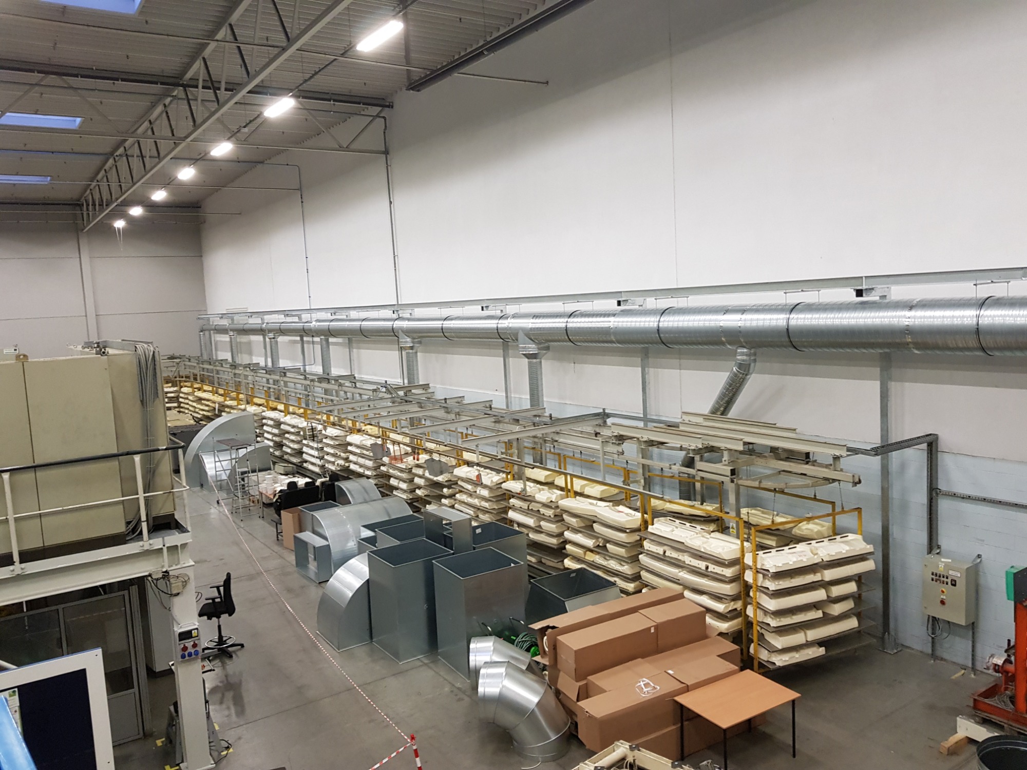 Wysoki kąt widzenia na magazyn z elementami instalacji wentylacyjnej, w tym rury, kanały i komponenty składowane na półkach, z widocznymi elementami systemu wentylacyjnego pod sufitem.