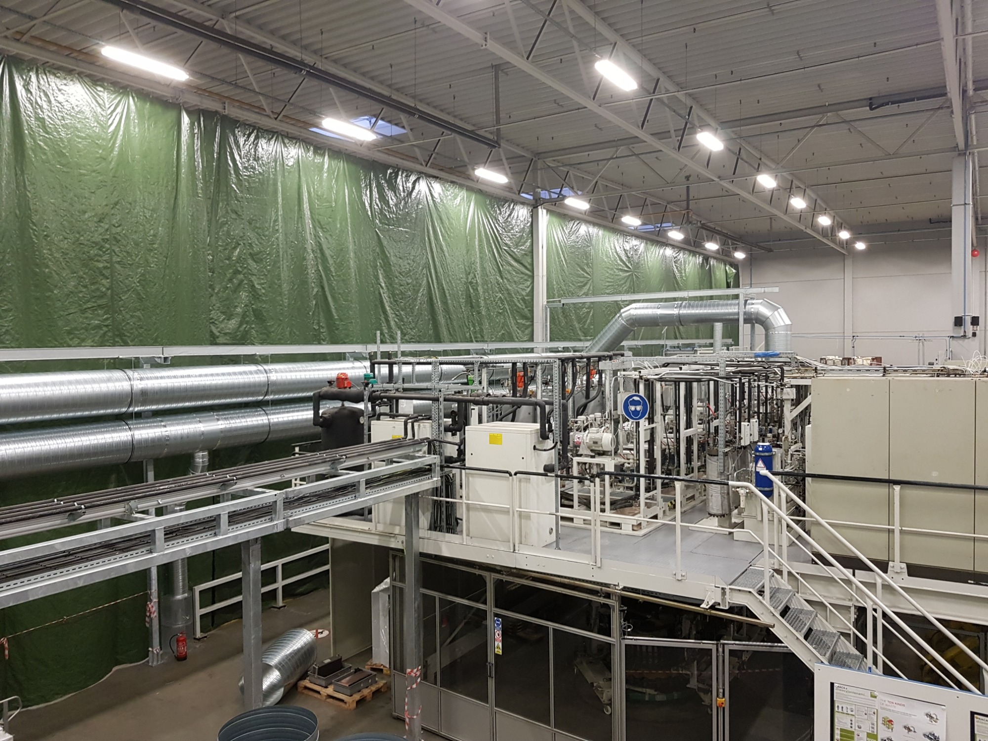 Widok z góry na instalację przemysłową z rurami, przewodami i urządzeniami na platformie, z zielonymi zasłonami w tle, oświetloną szeregiem lamp sufitowych.