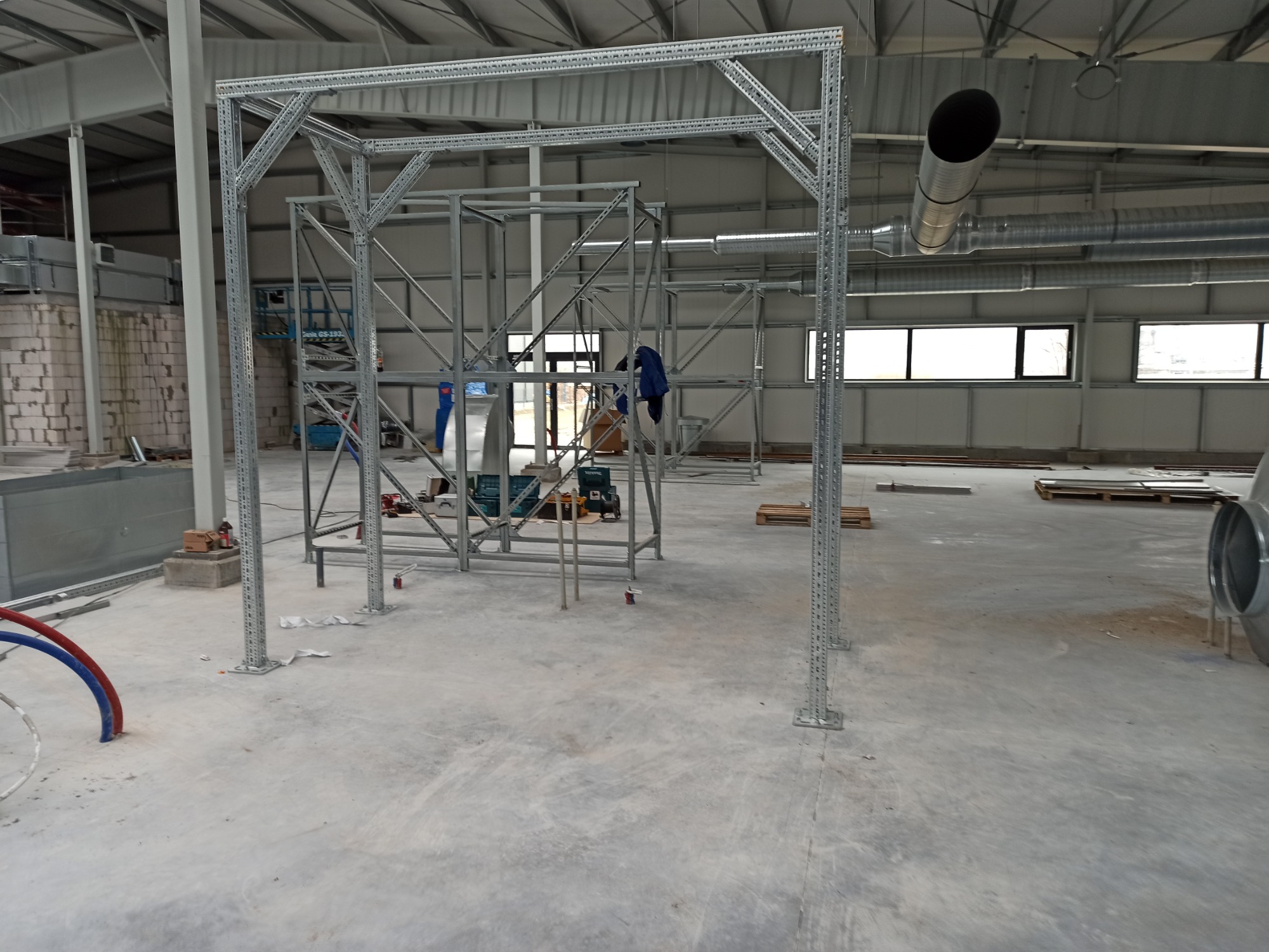 Wnętrze hali przemysłowej z widoczną konstrukcją wsporczą z profili perforowanych, prawdopodobnie pod system wentylacyjny, z pracownikami w tle i elementami instalacji na podłodze.