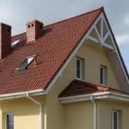 Żółta elewacja domu jednorodzinnego z czerwoną dachówką, białymi rynnami i kominem. Widoczne okna dachowe i trójkątny szczyt dachu z dekoracyjnymi elementami.