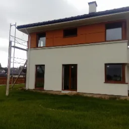 Dwukondygnacyjny dom z częściowo otynkowaną elewacją w kolorze złamanej bieli i panelami w odcieniach brązu, widoczne rusztowanie przy ścianie, trawnik przed domem, pochmurne niebo.