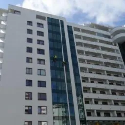 Dwóch alpinistów na szklanej fasadzie wysokiego, białego budynku z balkonami, pracujących na linach w słoneczny dzień.