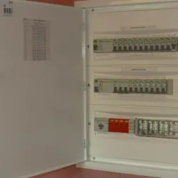 Otwarta biała skrzynka rozdzielcza z widocznymi bezpiecznikami i listwami zaciskowymi na tle surowych ścian.