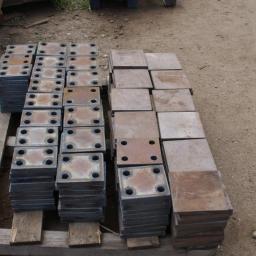 Anstal - Stosy metalowych, kwadratowych płyt z otworami, ułożone na drewnianej palecie na ziemi. Część płyt jest surowa, część pomalowana na ciemny kolor.