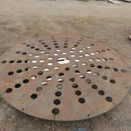 Anstal - Okrągły, metalowy element z regularnie rozmieszczonymi otworami, leżący na ziemi. Widoczne ślady obróbki i rdzawy nalot.