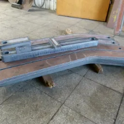 Metalowe elementy konstrukcyjne z otworami, ułożone warstwowo na podłodze w Będzinie. Widoczne ślady obróbki. Detal z bliska, surowy wygląd.