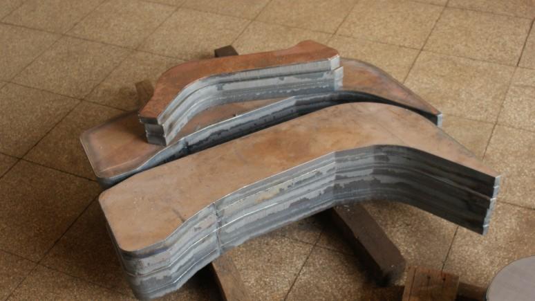 Stos wyciętych, metalowych elementów o nieregularnym kształcie, ułożonych warstwami na drewnianych podkładkach. Widoczne ślady obróbki i surowy wygląd metalu.