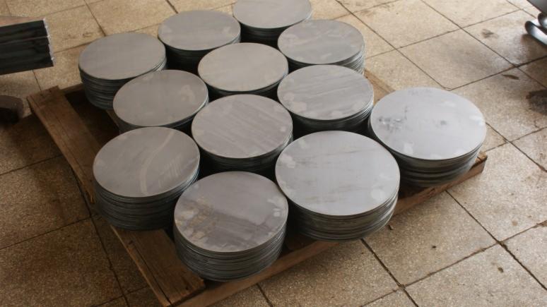 Stosy okrągłych, metalowych elementów ułożone na drewnianej palecie na tle płytek podłogowych. Elementy po obróbce, gotowe do dalszego wykorzystania.