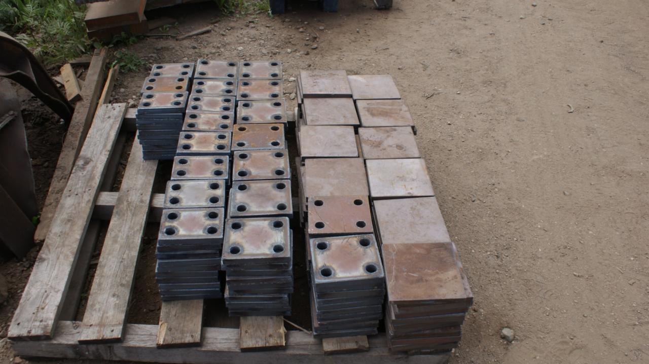 Stosy metalowych, kwadratowych płyt z otworami, ułożone na drewnianej palecie na ziemi. Część płyt jest surowa, część pomalowana na ciemny kolor.