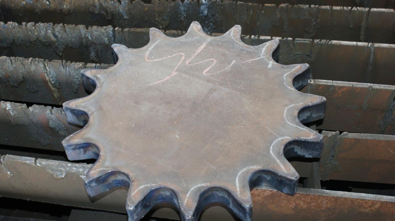 Wycięty z metalu element w kształcie koła zębatego, leżący na metalowej powierzchni z widocznymi śladami cięcia. Na elemencie widoczne delikatne, jasne oznaczenia.