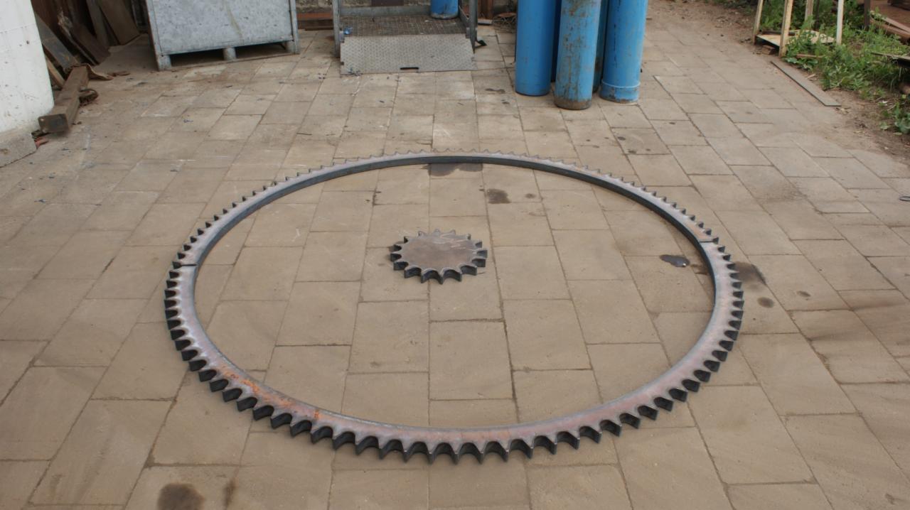 Dwa stalowe koła zębate, jedno duże w kształcie okręgu, drugie mniejsze, leżą na brukowanej powierzchni. W tle butle gazowe i metalowe konstrukcje.