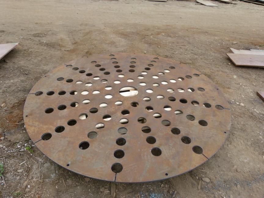 Okrągły, metalowy element z regularnie rozmieszczonymi otworami, leżący na ziemi. Widoczne ślady obróbki i rdzawy nalot.