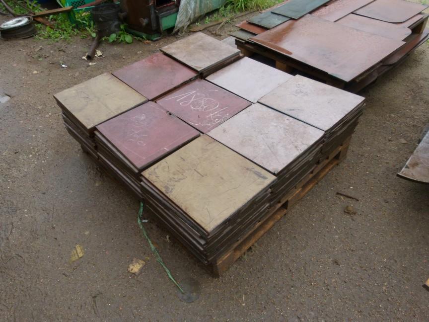 Stos kwadratowych blach na palecie, niektóre z napisami, na zewnątrz, obok leżą inne blachy.