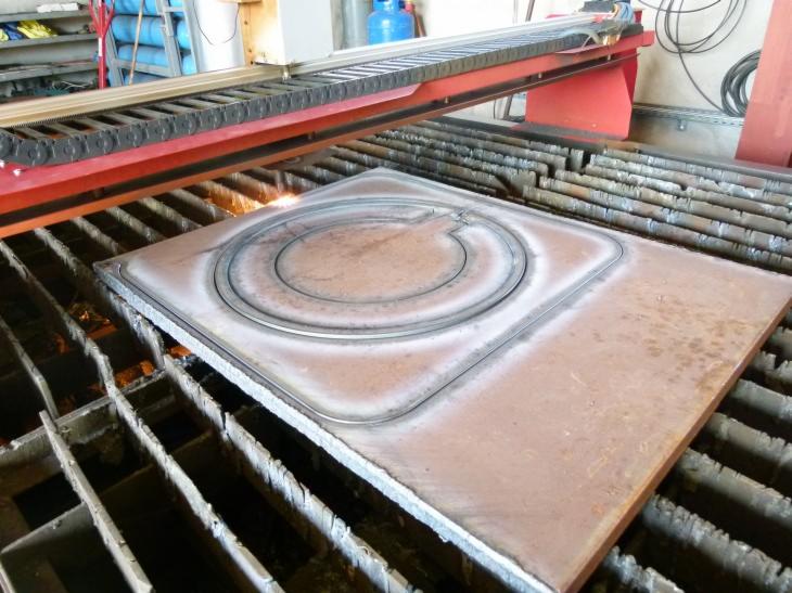 Proces wycinania plazmowego w metalu: kwadratowa płyta z wyciętym okręgiem i kształtem litery 'C', maszyna z czerwonymi elementami, iskry w miejscu cięcia.