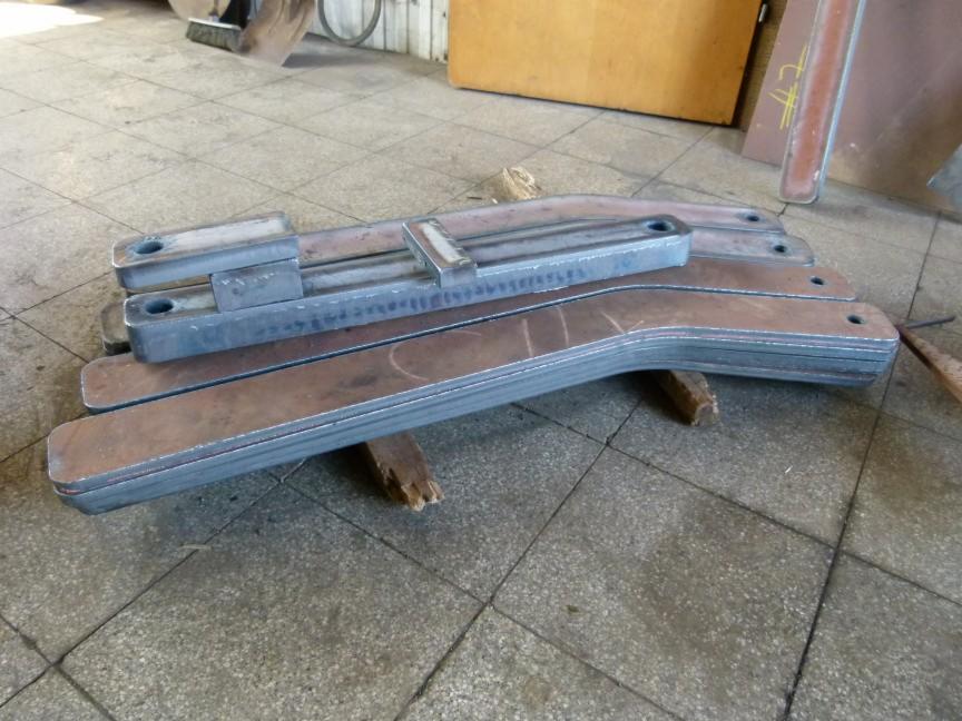 Metalowe elementy konstrukcyjne z otworami, ułożone warstwowo na podłodze w Będzinie. Widoczne ślady obróbki. Detal z bliska, surowy wygląd.