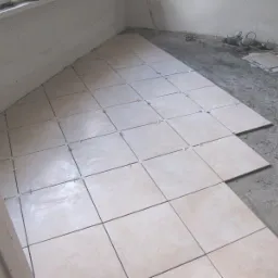 Układanie beżowych płytek podłogowych z krzyżykami dystansowymi w rogu pomieszczenia. Na podłodze widoczne narzędzia i nieułożone płytki.