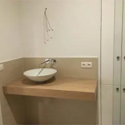 Nowoczesna łazienka z umywalką nablatową na drewnianym blacie i białymi szafkami z ciemnymi uchwytami, widoczne gniazdko elektryczne i włącznik światła.