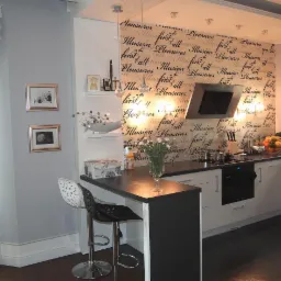 Nowoczesna kuchnia z wyspą i dwoma hokerami. Ściana zdobiona tapetą w napisy, na półkach dekoracje. Białe meble i ciemny blat. Oświetlenie punktowe i wiszące lampy.