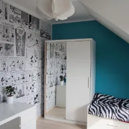 Pokój dziecięcy na poddaszu: białe meble, komiksowe tapety i turkusowa ściana. Lustrzane drzwi szafy odbijają wnętrze, a lampa sufitowa dodaje uroku.