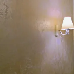 Ściana z dekoracyjną fakturą w odcieniach beżu i brązu, oświetlona kinkietem z białym abażurem i kryształkami.