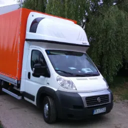 Biały bus z pomarańczową plandeką na tle wiejskiego krajobrazu. Widoczne krzewy róż i budynek gospodarczy. Tablica rejestracyjna z przodu pojazdu.