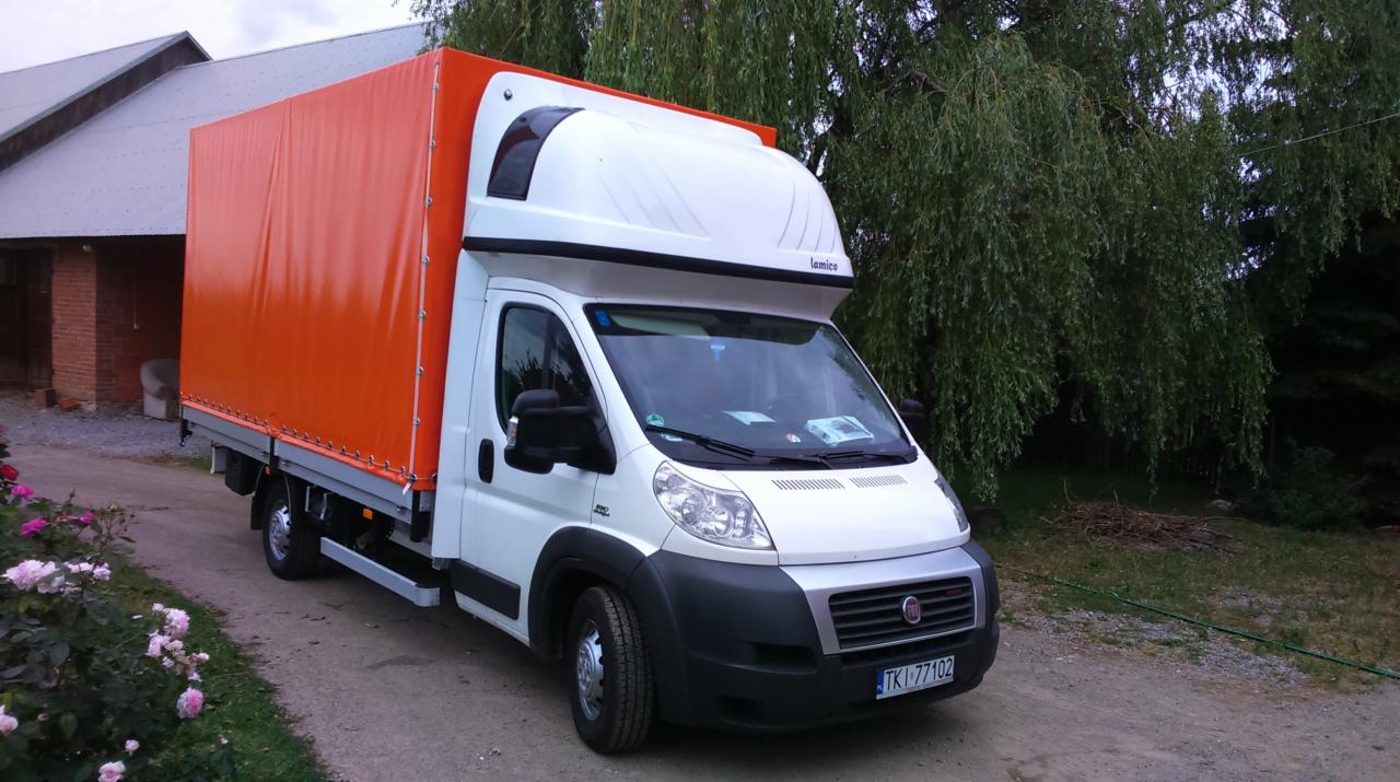 Biały bus z pomarańczową plandeką na tle wiejskiego krajobrazu. Widoczne krzewy róż i budynek gospodarczy. Tablica rejestracyjna z przodu pojazdu.