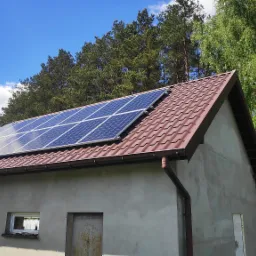 Panele fotowoltaiczne na brązowym dachu budynku gospodarczego, widok z boku na tle drzew i błękitnego nieba z chmurami.