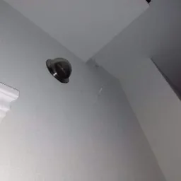 Świeżo zamontowany, okrągły anemostat wentylacyjny z metalową obudową, umieszczony na jasnoszarej ścianie w rogu pomieszczenia, widoczny fragment listwy przypodłogowej.