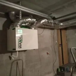 Zamontowany rekuperator marki Wanas z podłączonymi przewodami wentylacyjnymi w surowym pomieszczeniu z betonową ścianą i drabinką.