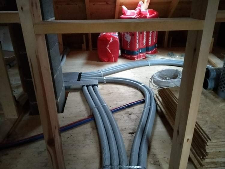 Szare, karbowane rury wentylacyjne układane w drewnianej konstrukcji poddasza w Ostrołęce, widoczne czerwone paczki izolacji Rockwool w tle.