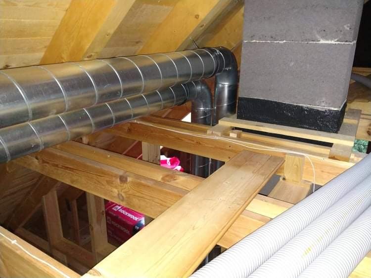 Dwie metalowe rury wentylacyjne spiralne, połączone kolankami, prowadzone w przestrzeni poddasza o drewnianej konstrukcji, obok komina z szarych bloczków, z widoczną izolacją Rockwool.