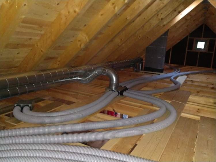Instalacja systemu wentylacji z szarymi elastycznymi rurami w przestrzeni poddasza wykończonego drewnem, widoczne metalowe kanały wentylacyjne i czarny komin.