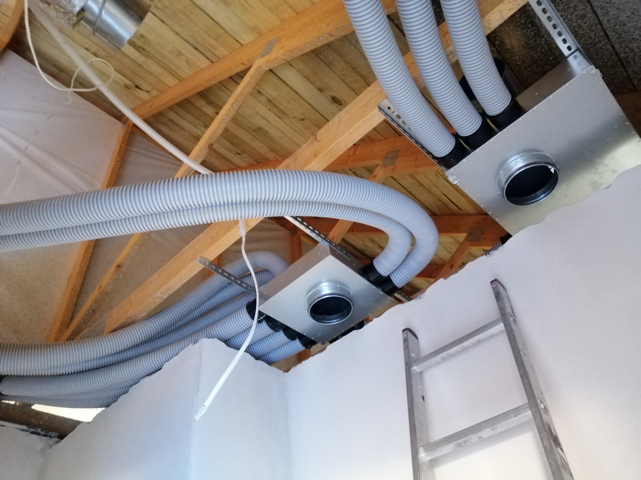 Szare, elastyczne przewody wentylacyjne podłączone do metalowych skrzynek rozdzielczych w drewnianym stropie, widoczne elementy izolacji i drabina oparta o białą ścianę.
