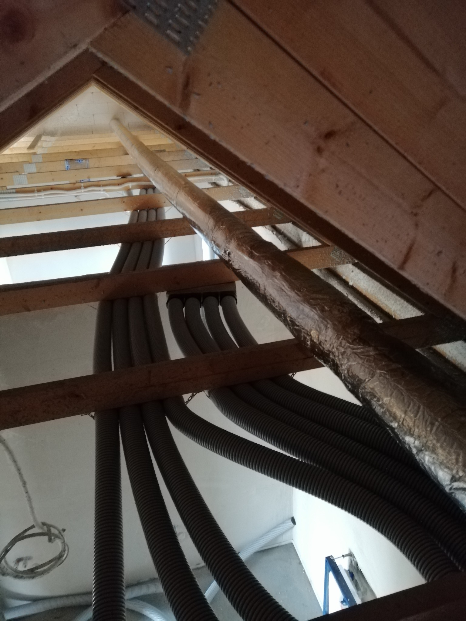 Szare, karbowane rury wentylacyjne rozprowadzone pod drewnianą konstrukcją dachu na poddaszu, widoczny fragment rury owiniętej srebrną folią izolacyjną.