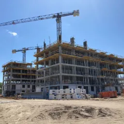 Wysoki budynek w trakcie budowy z widocznymi żelbetowymi konstrukcjami, drewnianym szalunkiem i dwoma żurawiami na tle błękitnego nieba.