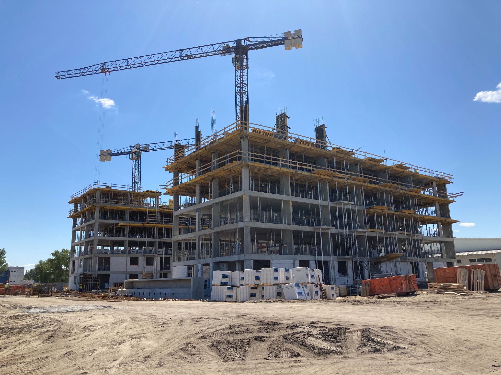 Wysoki budynek w trakcie budowy z widocznymi żelbetowymi konstrukcjami, drewnianym szalunkiem i dwoma żurawiami na tle błękitnego nieba.