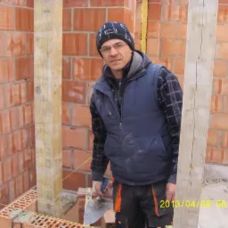 Murarz w czapce i okularach, z kielnią w dłoni, pozuje na tle wznoszonej ściany z czerwonej cegły i drewnianych słupów, z datą wykonania zdjęcia w prawym dolnym rogu.