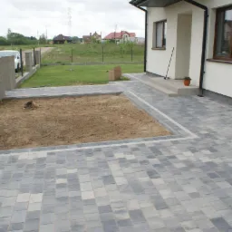 Ułożona szara nawierzchnia z kostki, obrzeże z jasnej kostki, przygotowany teren pod trawnik, w tle dom jednorodzinny i ogrodzenie posesji.