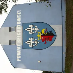 Ściana budynku z herbem Powiatu Bełchatowskiego, namalowanym na elewacji w kolorach niebieskim, białym, żółtym, czerwonym i czarnym.