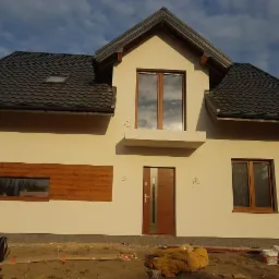 Dwukondygnacyjny dom z beżową elewacją, brązowymi oknami i drzwiami, oraz ciemnoszarą dachówką, z widocznymi śladami prac wykończeniowych wokół budynku.