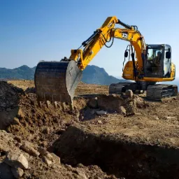 Żółta koparka JCB wykonuje wykop w ziemi na tle gór i morza, widok z dołu.