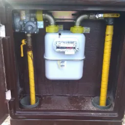 Zewnętrzna szafka gazowa z widocznym licznikiem gazu, rurami i zaworami, żółte rury zabezpieczone taśmą, brązowa szafka z otwartymi drzwiczkami.